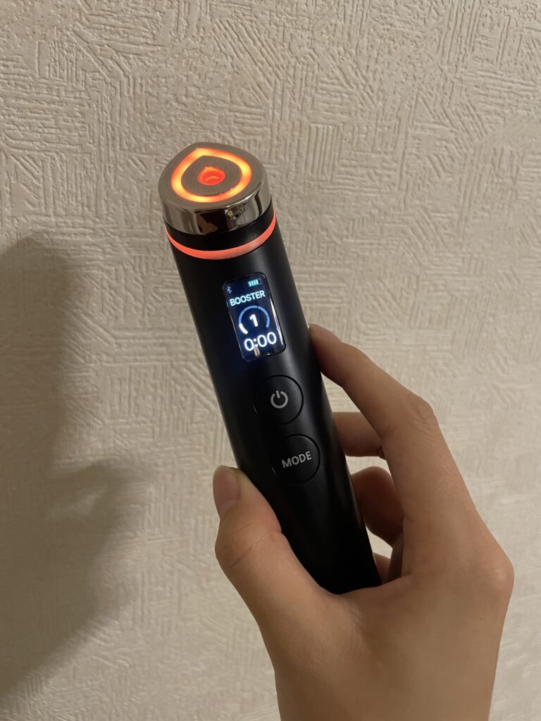 メディキューブの美顔器「AGE-Rブースタープロ」の実物を手に持ち、液晶画面（LCD）と赤色LEDリングの発光状態を示した写真